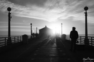 Black and white silhouette of a person walking along Manhattan Beach Pier at sunset