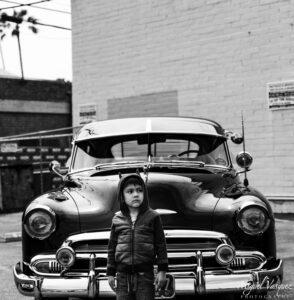 Black and white portrait of a young boy standing beside a classic lowrider car in Santa Monica